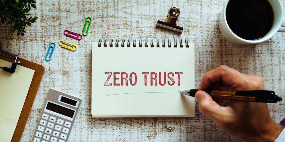 Am image of a desk containing a coffee mug, paper clips, magnetic clip, clipboard with paper, a calculator, and a notebook with the words "ZERO TRUST" stamped in red on it and someone is under lining it.