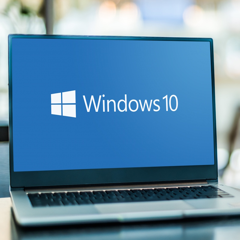 Windows 10 Laptop with a blue screen background with white text that says "Windows 10". This is on a silver laptop with a blurry background behind it.