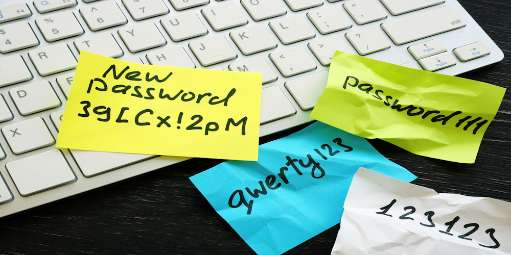 Sitcky notes with crumbled passwords on them reading, "New Password 3g[Cx!2pM", "Password111", "qwerty123", and "123123". Behind this is a white keyboard on a black wooden table.