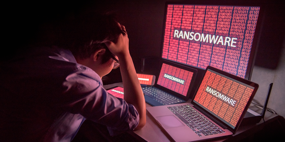 A person sits at a desk with their head in their hand, surrounded by multiple computer screens displaying the word "RANSOMWARE" in large letters over red backgrounds filled with binary code.